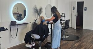 A hairstylist wearing gloves works on a seated client's hair in a modern, well-lit salon with wooden floors and round mirrors on the wall. | In Bloom Salon & Spa, Monterey CA